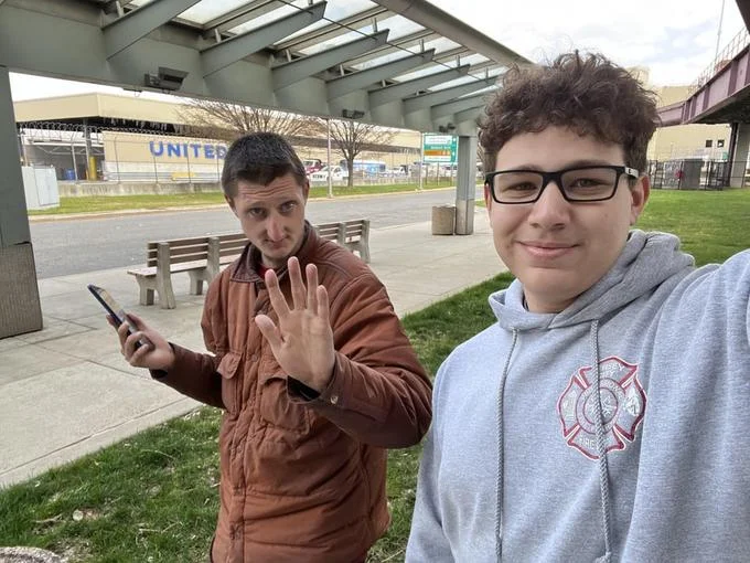 Daniel Larson X Joshua Block World Of T Shirts — Daniel Larson waving outdoors while a fan takes a selfie with him near a bench