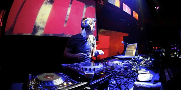 Darude Sandstorm — Performance photograph of DJ Darude actively DJing at turntables on concert stage with dramatic stage lighting.