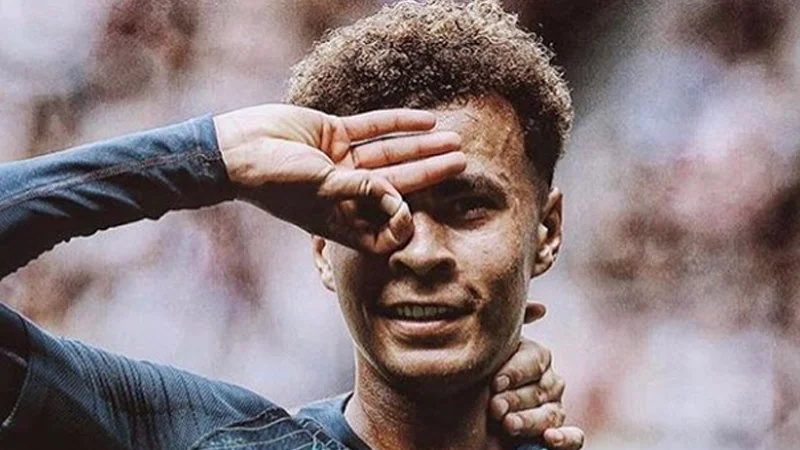 Dele Alli Hand Celebration — Professional close-up photograph of Dele Alli in Tottenham kit performing signature hand gesture with OK sign to eye.