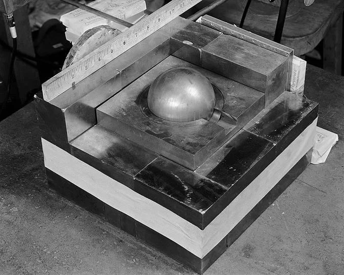 Black-and-white photo of the demon core plutonium sphere partially surrounded by stacked tungsten carbide reflector blocks.
