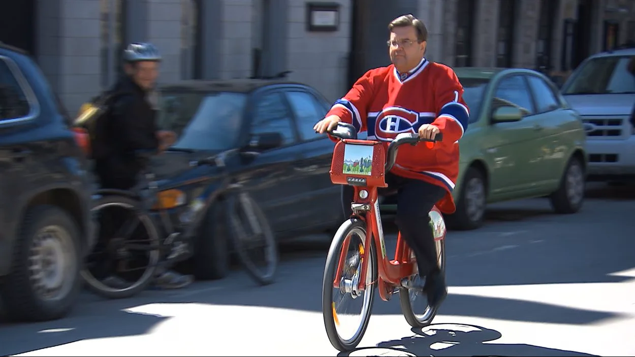 Denis Coderre riding a Bixi — Denis Coderre in a red Canadiens jersey riding a red Bixi bike on a Montreal street