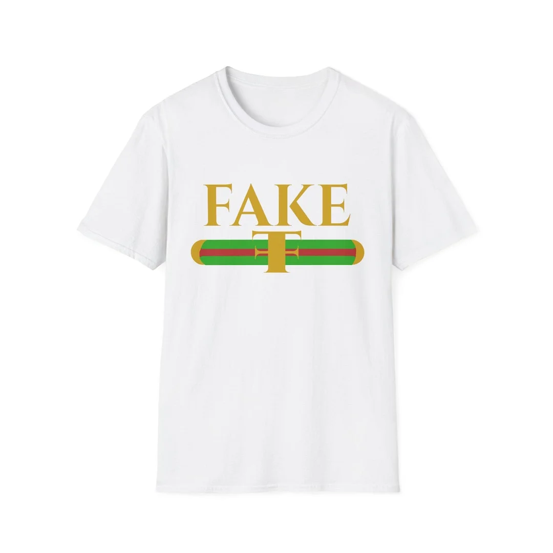 Designer Knockoff — White t-shirt featuring prominent gold 'FAKE' text with red and green horizontal stripe pattern parodying Gucci luxury brand.
