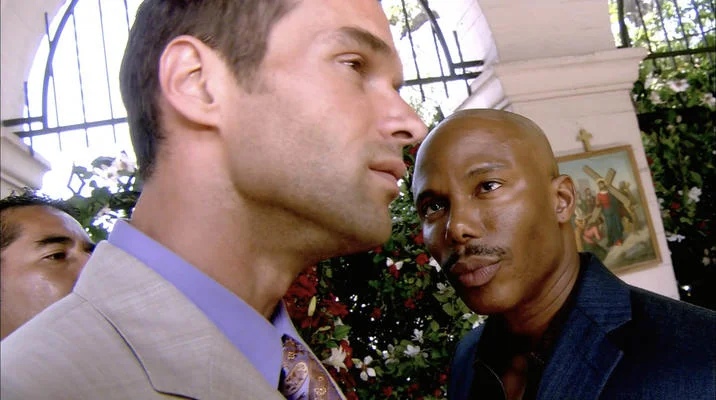 Doakes Looking at Guerrero — Wider Dexter screenshot of Doakes in profile staring at a bald man in a dark suit outside a small shrine.