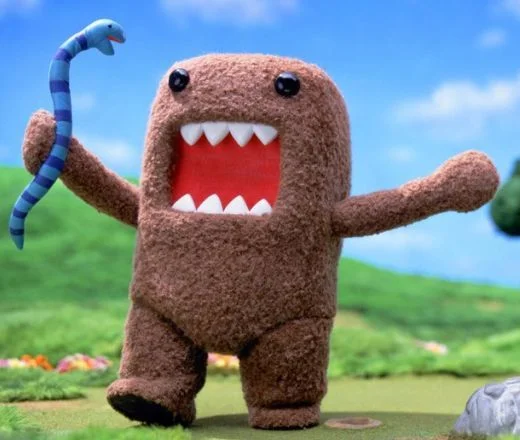 Domo-kun brown furry mascot with open toothy mouth holding a blue snake in a grassy field