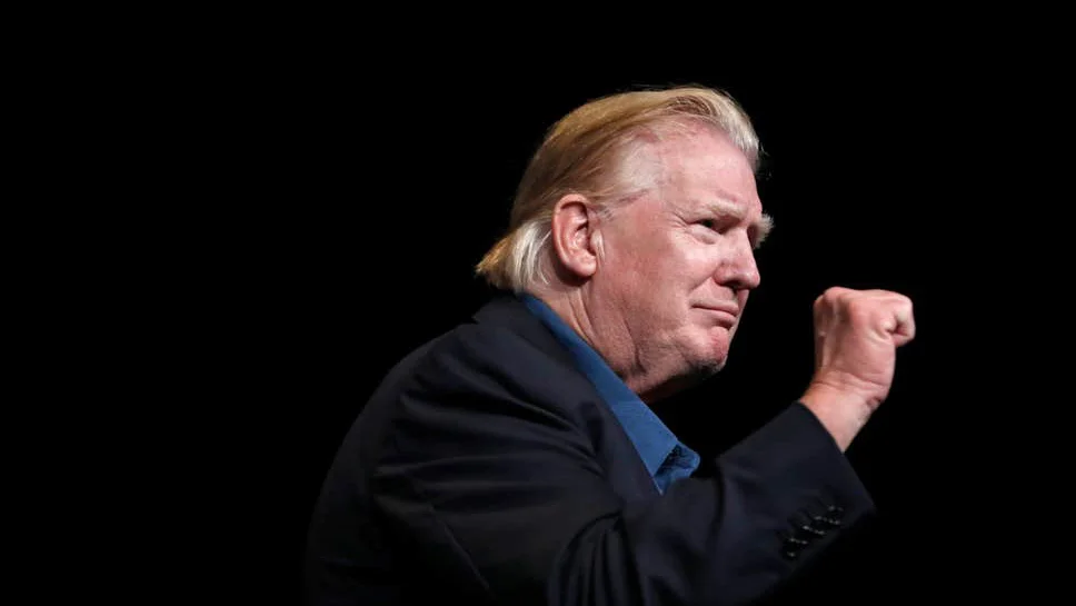 Donald Trumps Slicked Back Hair — Donald Trump in profile with slicked-back hair raising a fist on a dark stage