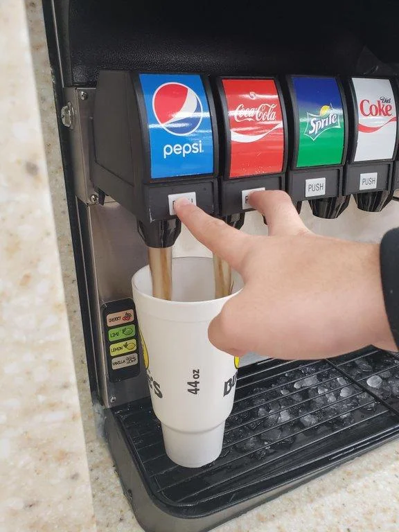Double Soda Pour — Vertical photo of a hand pressing Pepsi and Coca-Cola dispenser buttons at the same time into a 44 oz cup.