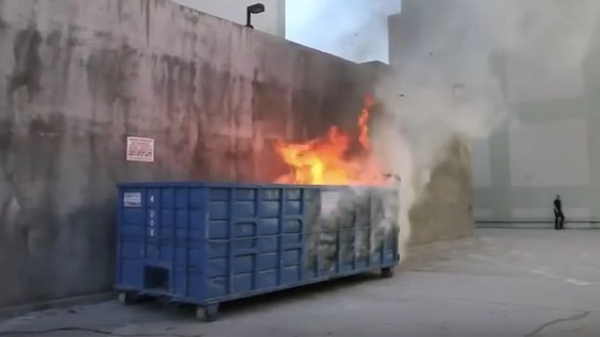 Dumpster Fire — Blue industrial dumpster on fire against a concrete wall with smoke rising
