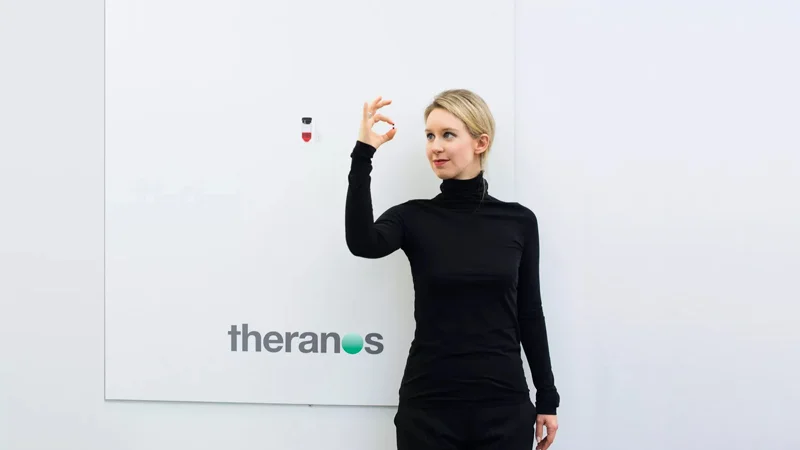 Elizabeth Holmes Theranos Fraud Case — Elizabeth Holmes in black turtleneck holding a Theranos nanotainer blood vial in front of the company logo