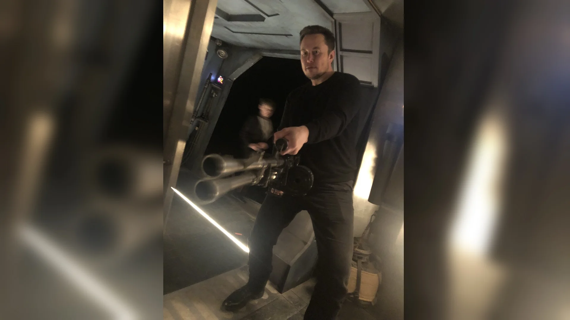 Elon Musk Holding A Minigun — Elon Musk holding a large multi-barreled minigun prop weapon in an industrial setting