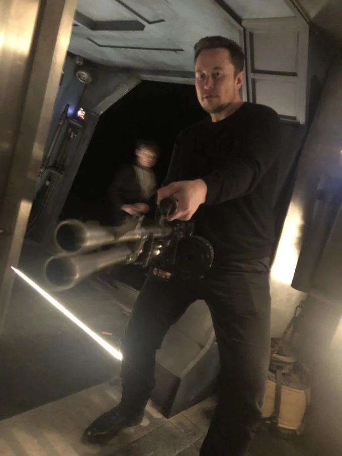 Elon Musk Holding A Minigun — Vertical photo of Elon Musk aiming a minigun prop weapon in a dark corridor