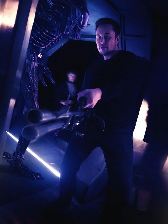 Elon Musk Holding A Minigun — Photoshop edit of Elon Musk with minigun and a Xenomorph alien in the background