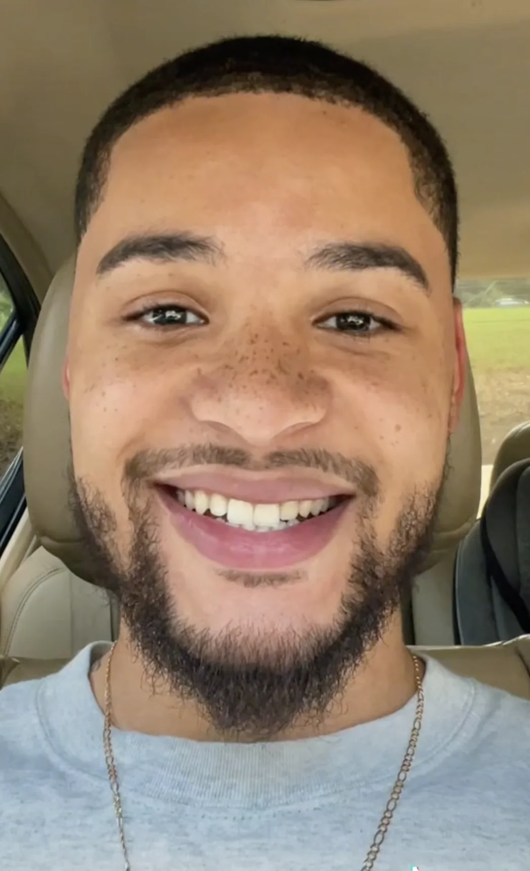 Enoch J TikTok Duets — Close-up selfie of Enoch J smiling broadly in a car wearing a gray shirt and gold chain.