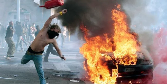Er Pelliccia — Shirtless man throwing red fire extinguisher at flaming barricade during urban protest.