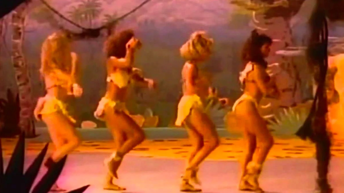 Everybody Walk The Dinosaur — Four women in cavewoman costumes dancing on a prehistoric-themed stage from the Walk the Dinosaur music video