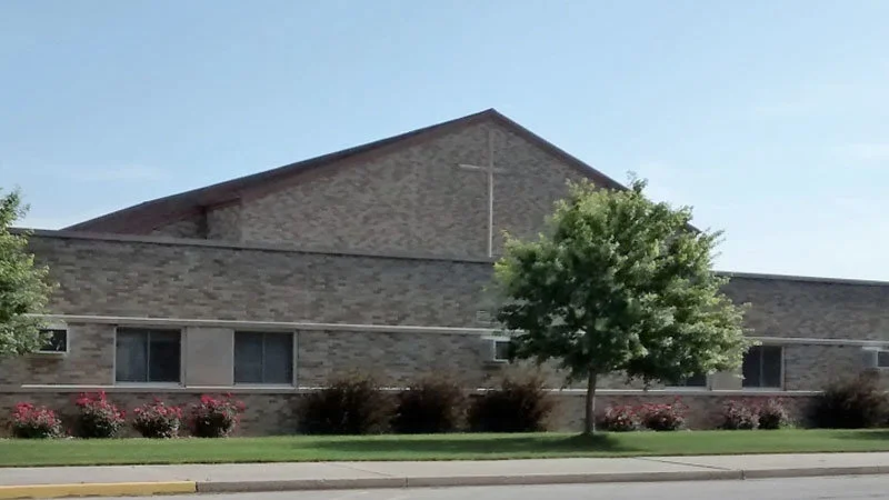 Exposechristianschools — Low-rise brick building with prominent cross on peaked roofline surrounded by trees.