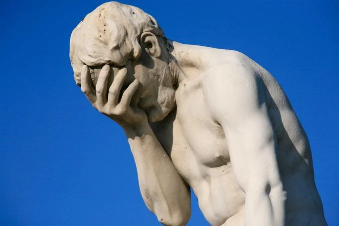Facepalm — White marble statue of hunched figure pressing hand to face against blue sky.