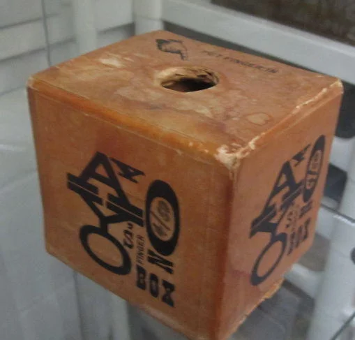 Finger Boxes — Aged orange AY-O Finger Box cube in a museum vitrine with 'put finger in' label.