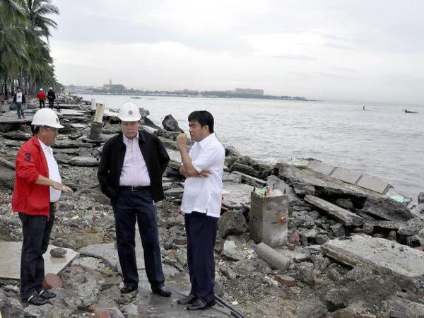 Floating Filipino Government Officials — Three men in hard hats at rocky beach with typhoon debris and palm trees.