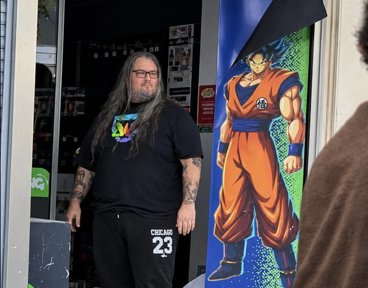 French Goku Hero photo: long-haired bearded man in black T-shirt stands in shop doorway beside life-size Goku banner.