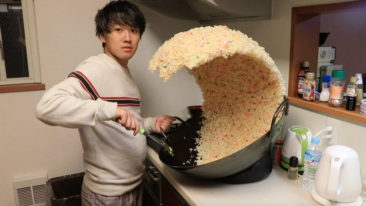 Fried Rice Prank — Man in kitchen holding spatula next to a wok with a giant wave of fried rice