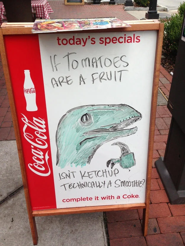 Funny Store Signs — Sidewalk chalkboard sign joking that ketchup is technically a smoothie, with a doodled dinosaur