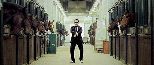PSY dressed in a formal tuxedo performing the horse-riding Gangnam Style dance move in a stable surrounded by horses.