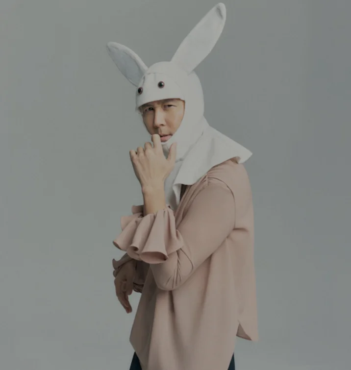 Gi Hun Bunny — Professional studio photograph of Lee Jung-jae in white bunny costume with raised hand in contemplative gesture against gray background.
