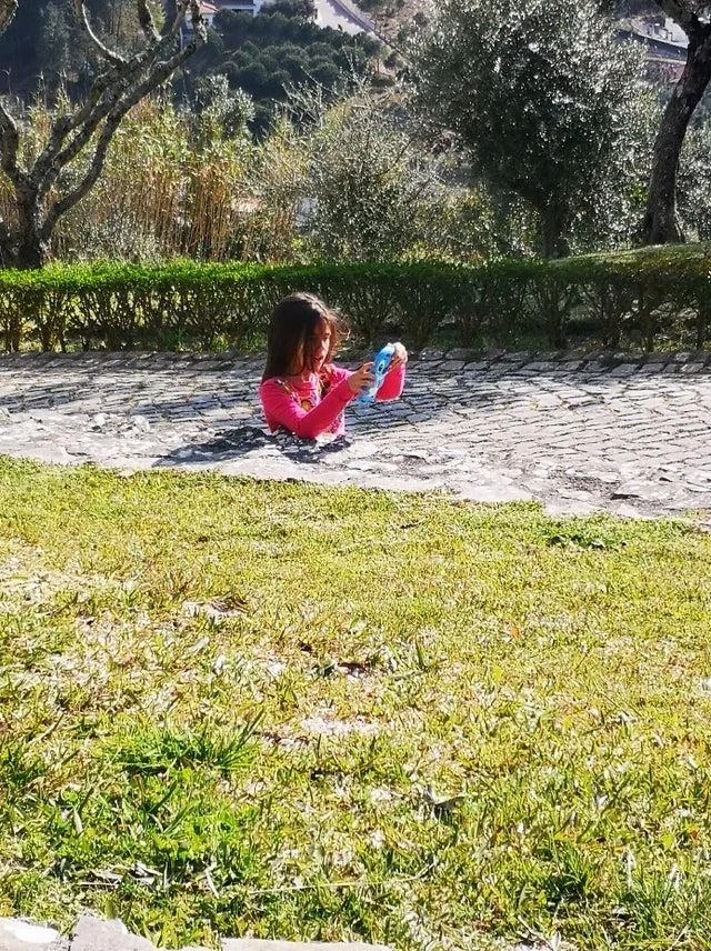 Girl Stuck In Concrete Illusion — A young girl in pink top sits on a stone path with grass foreground at same visual level, making her lower body appear