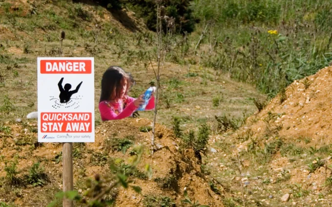 Girl Stuck In Concrete Illusion — Girl in pink composited with red-and-white 'DANGER QUICKSAND STAY AWAY' warning sign in dirt hillside setting.