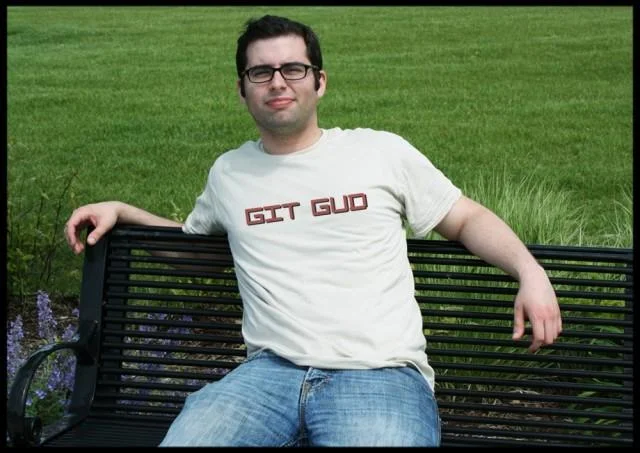 Young man with glasses sitting on park bench wearing white t-shirt with 'GIT GUD' in red letters.