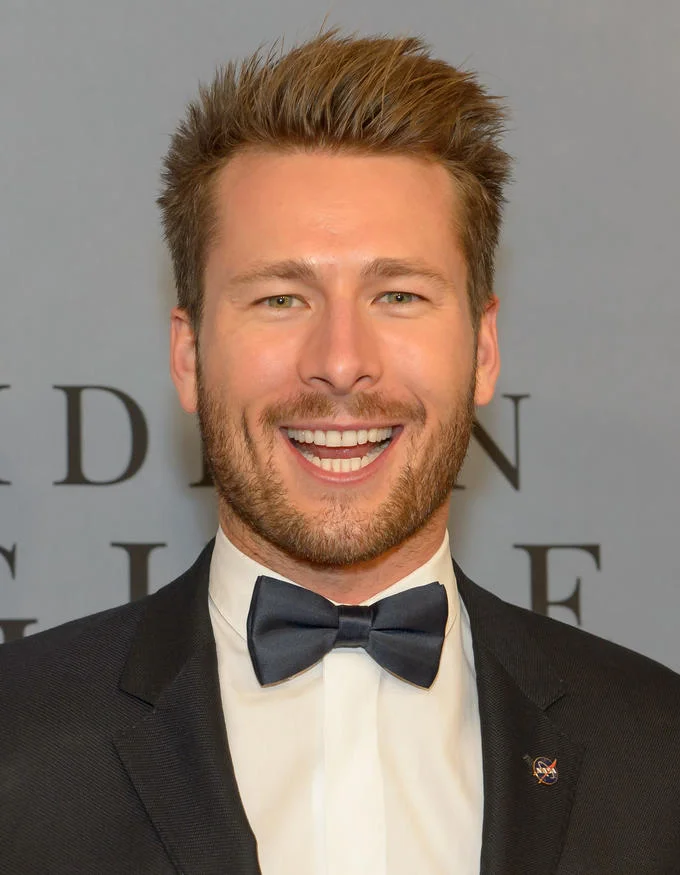 Glen Powell Capybara Face — Vertical portrait of Glen Powell smiling broadly in a tuxedo with a NASA pin on his lapel.