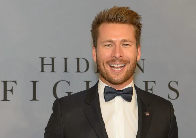 Glen Powell Capybara Face — Glen Powell smiling in a black tuxedo in front of a Hidden Figures premiere backdrop.