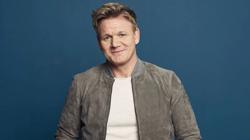 Gordon Ramsay smirking in grey jacket against blue background