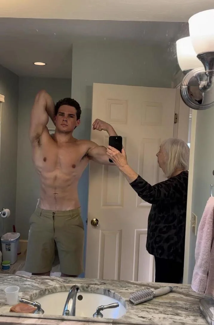 Grandma Taking Photo of Grandson In Bathroom Mirror — Shirtless young man flexing bicep as grandmother photographs him in bathroom mirror.