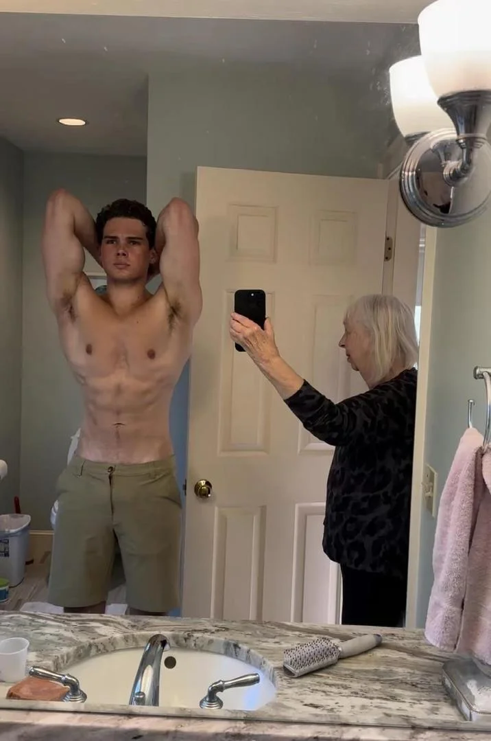 Grandma Taking Photo of Grandson In Bathroom Mirror — Shirtless young man flexing in bathroom mirror while grandmother takes his photo with a phone.