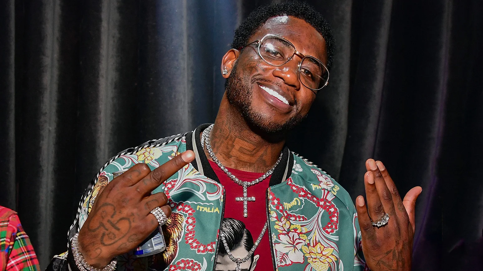 Gucci Mane smiling and flashing a peace sign while wearing a floral Gucci jacket and diamond chain