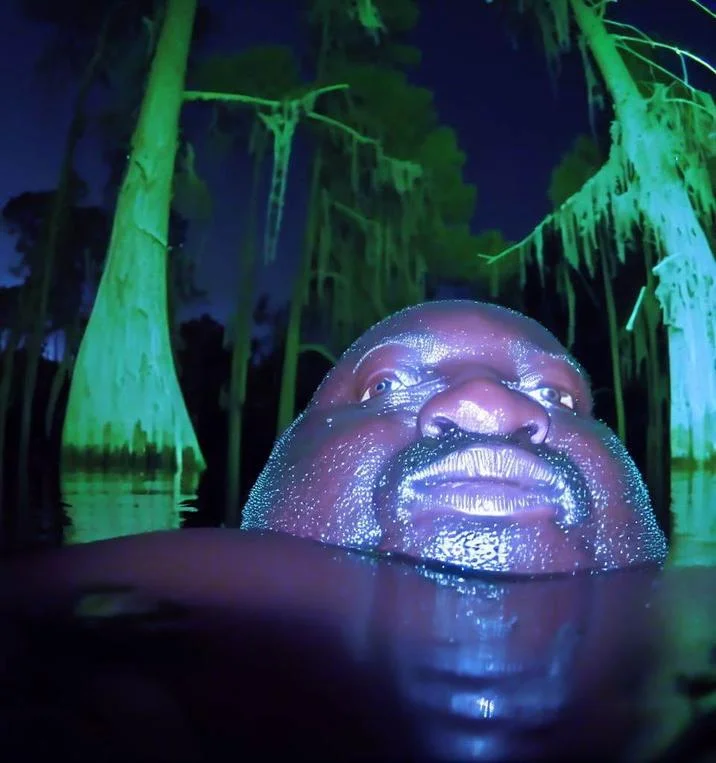 Gumbo Slice — Man's head emerging from dark swamp water surrounded by cypress trees at night