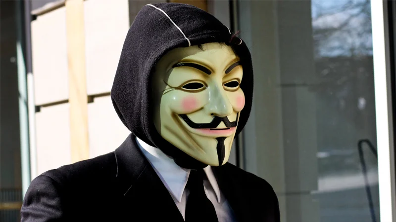 Person in suit and hood wearing a Guy Fawkes mask