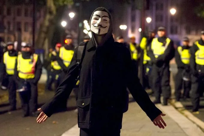 Protester in Guy Fawkes mask facing riot police at night