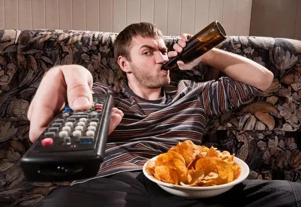 Guy Sipping Beer On A Couch Stock Image — Original stock photo of man in striped shirt on floral couch, sipping beer bottle while pointing TV remote at camera.