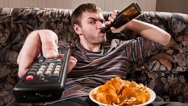 Guy Sipping Beer On A Couch Stock Image — Canonical stock photo of man in striped shirt guzzling beer on floral couch with remote and chips.