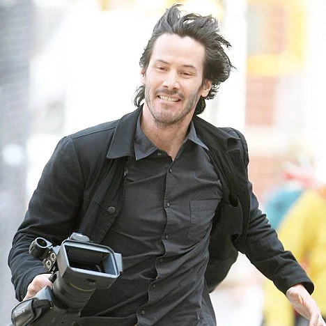 Happy Keanu — Keanu Reeves smiling and running down a street holding a camera, wearing a black jacket and shirt
