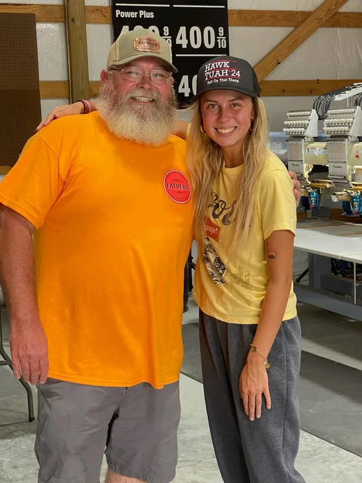 Hawk Tuah Girl — Hailey Welch in a Hawk Tuah 24 hat posing with a bearded man at a merch shop