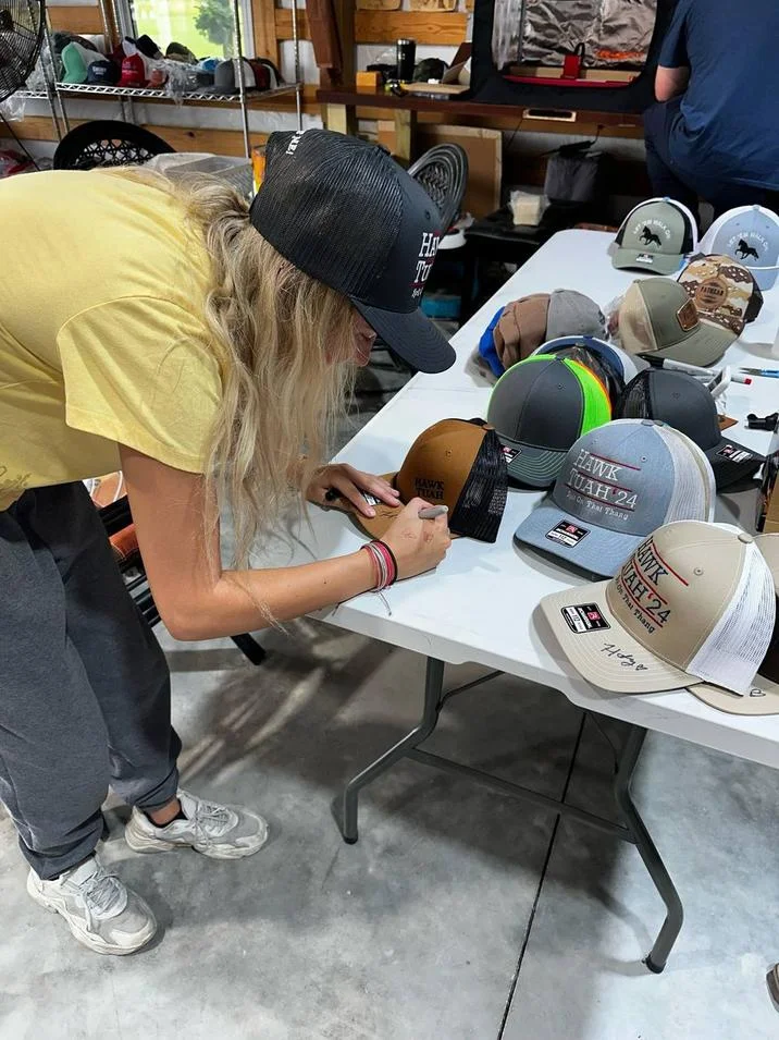 Hawk Tuah Girl — Hailey Welch signing autographs on Hawk Tuah trucker hats spread across a table