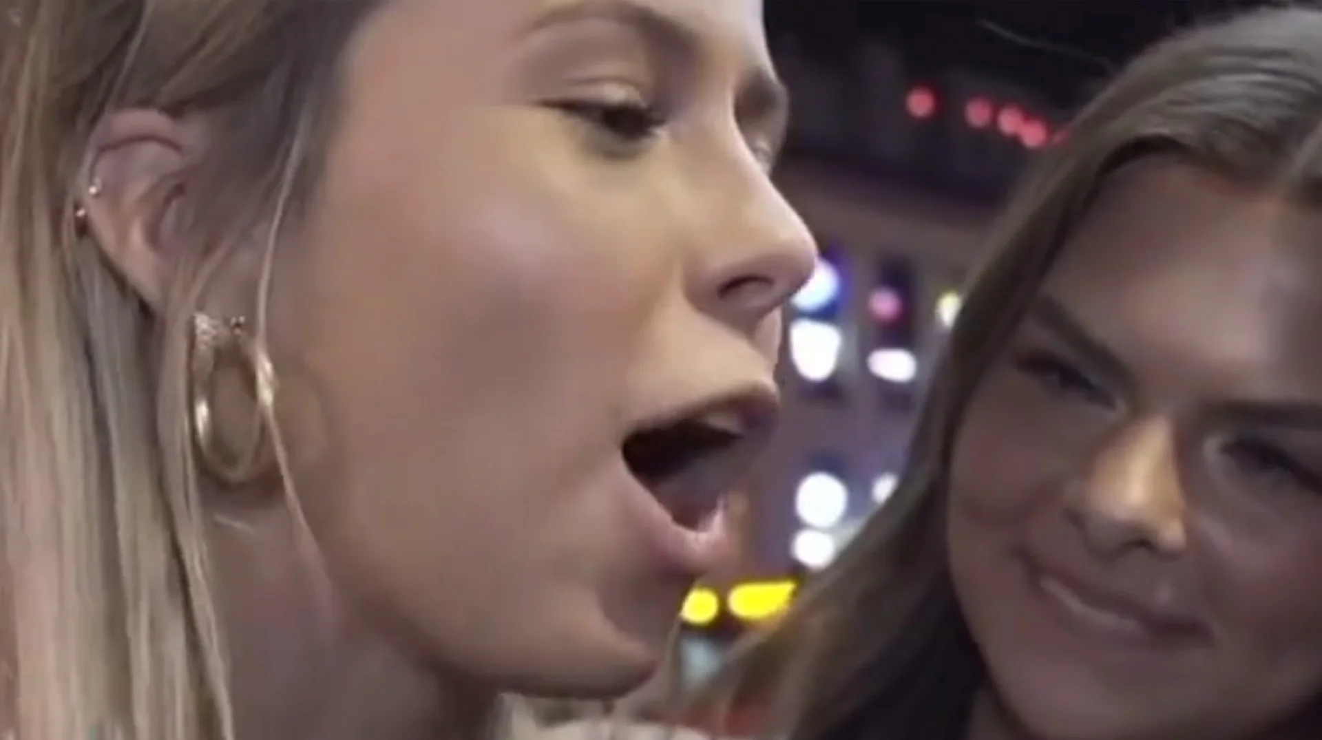 Hawk Tuah Girl — Blonde woman in profile with mouth open speaking during a night street interview, two friends beside her