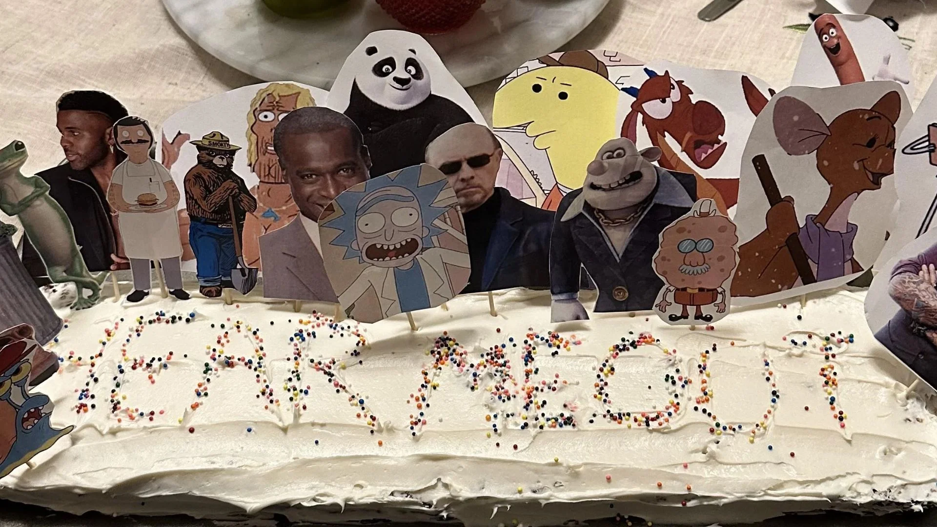 Hear Me Out Cake Trend — Frosted cake reading 'Hear Me Out' with paper cutouts of cartoon characters and celebrities stuck on top as toppers