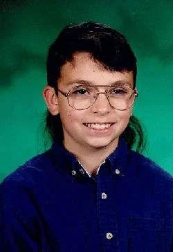 Hello My Future Girlfriend — A classic school portrait of a young boy wearing large wire-framed glasses and a blue button-down shirt against a green studio