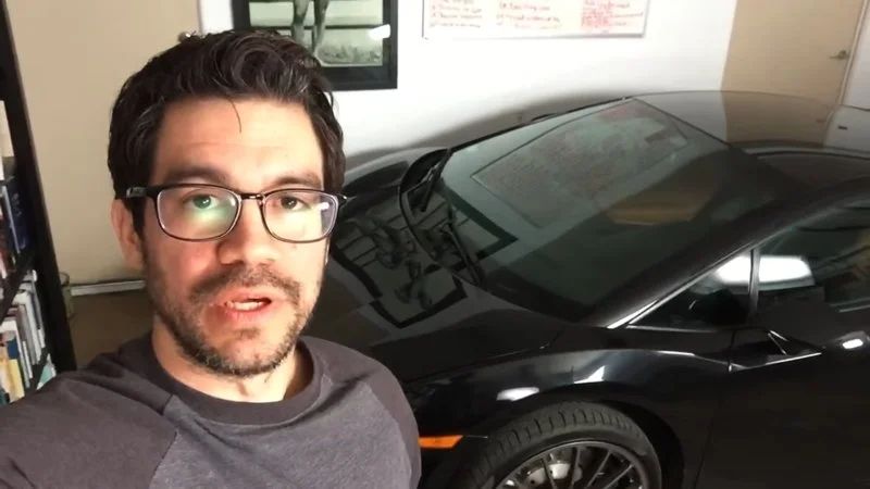 Here In My Garage — A man with glasses poses in front of an orange and dark sports car inside a garage with shelving and equipment visible in background.