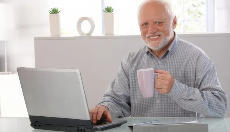 Hide the Pain Harold — The canonical stock photo showing András Arató at laptop in sunlit interior holding coffee mug with wide strained smile masking inner