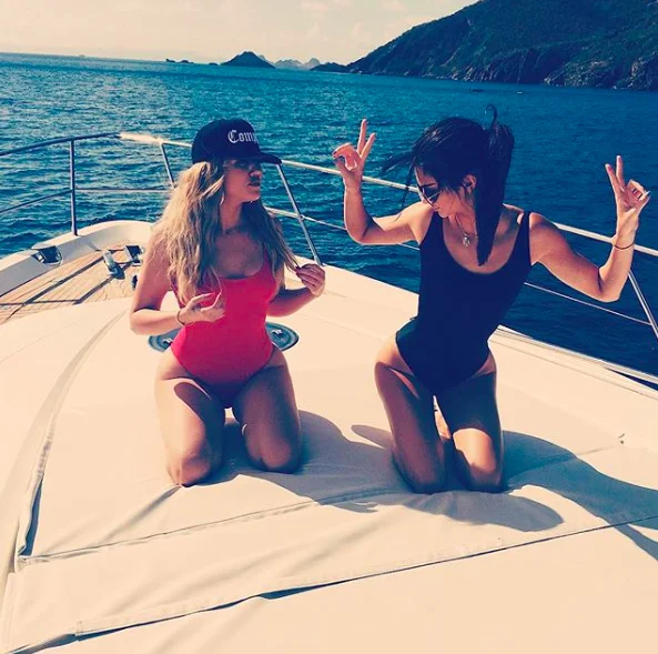 Hip Cleavage — Kardashian and Jenner in high-cut swimsuits kneeling on yacht deck with ocean backdrop